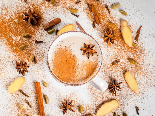 Collombatti Naturals 8 Reasons to Love Tea this Winter Blog Post Cover, A cup of chai on a white background with cinnamon sprinkled all around the cup with star anise, cloves, ginger and cinnamon sticks scattered around the cup
