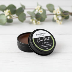 Blossom and Hive Choc-Mint lip balm  in black aluminium tin, sitting on a white painted timber surface with blurred greenery in the background