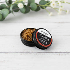 Blossom and Hive Honey Buff lip scrub in a black aluminium tin, sitting on a white painted timber surface with blurred greenery in the background
