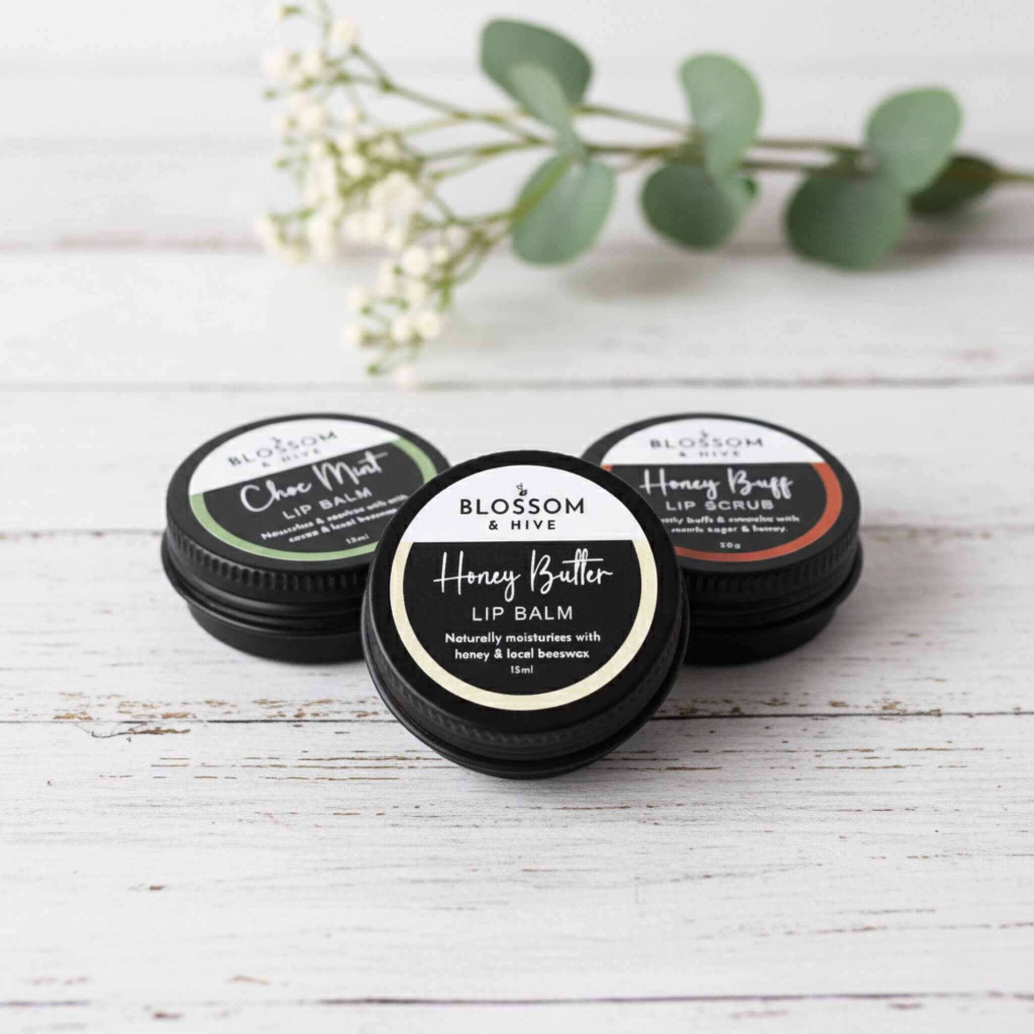 Blossom and Hive lip balm and lip scrub trio in black aluminium tins, sitting on a white painted timber surface with blurred greenery in the background
