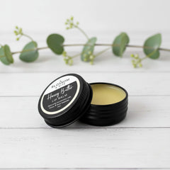 Blossom and Hive Honey lip balm in ablack aluminium tin, sitting on a white painted timber surface with blurred greenery in the background