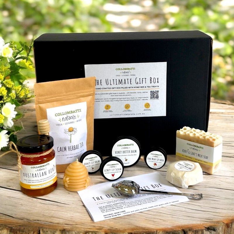 A gift box from Collombatti Naturals labelled "The Ultimate Gift Box" is displayed on a wooden surface with a green garden background. In front of the box are various handcrafted products including a pouch of Calm Herbal Tea, a jar of Australian honey with a wooden honey dipper, a beeswax candle shaped like a hive, several lip balms and skincare tins, a bar of goat’s milk soap, a tea infuser, and an information card. The scene is styled with fresh flowers, highlighting the natural, eco-friendly products