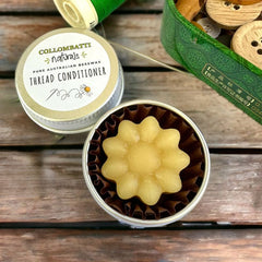 Collombatti Naturals Beeswax Thread Conditioner in a reusable tin. The thread wax, shaped like a delicate flower, sits in a paper liner for easy removal. Surrounded by sewing supplies, including spools of green thread and a vintage tin of wooden buttons, this all-natural beeswax thread conditioner is perfect for hand-stitching, embroidery, and quilting