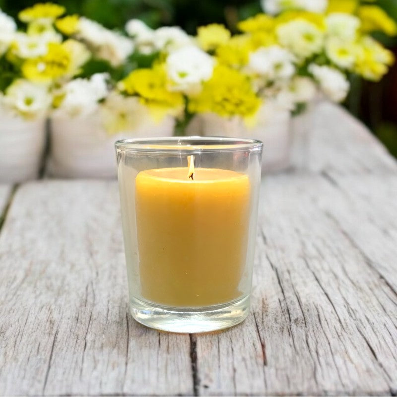 Beeswax votive candle burning in a clear glass holder on a rustic wooden surface, with blurred yellow and white flowers in the background.