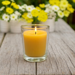 Beeswax votive candle burning in a clear glass holder on a rustic wooden surface, with blurred yellow and white flowers in the background.
