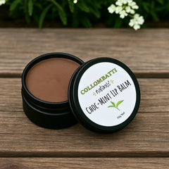 A tin of Collombatti Naturals Choc-Peppermint and Raw Honey Lip Balm, placed on a rustic wooden surface with fresh greenery and flowers in the background. The open tin reveals a smooth, rich brown balm, and the eco-friendly metal packaging features a white label with green and black branding, along with an illustration of a mint leaf, highlighting the natural ingredients.