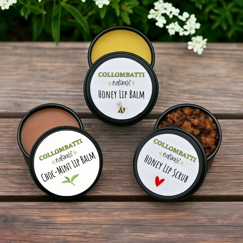 Three tins of Collombatti Naturals lip care products displayed on a rustic wooden surface with greenery and white flowers in the background. The open tins reveal a honey lip balm with Australian beeswax, a choc-peppermint and raw honey lip balm, and a honey lip scrub with a rich brown sugar texture. The eco-friendly metal tins feature white labels with green and black branding.