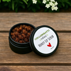 Collombatti Naturals Honey Lip Scrub in a round, reusable tin, placed on a rustic wooden surface with fresh white flowers in the background. The scrub has a rich, textured appearance, made with natural honey and sugar to soften and rejuvenate lips. The tin features a minimalist label with a heart symbol, reflecting the brand's eco-friendly and handmade ethos.