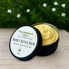 An open tin of Collombatti Naturals Honey Butter Balm, a rich, all-natural balm designed for very dry skin. The smooth, creamy texture of the balm is visible inside the tin, placed on a rustic wooden surface with fresh flowers in the background.